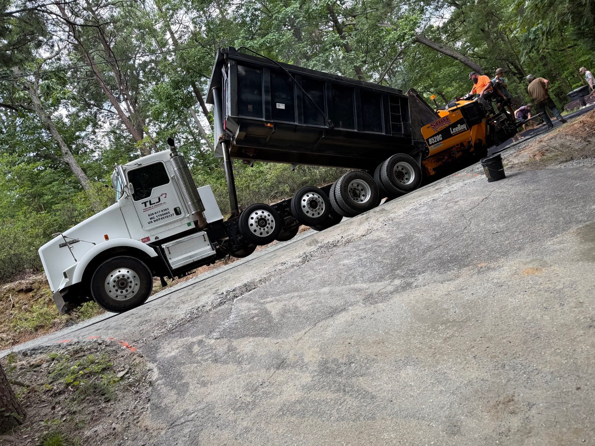 TLJ dump truck feeding an asphalt paver on a wooded driveway